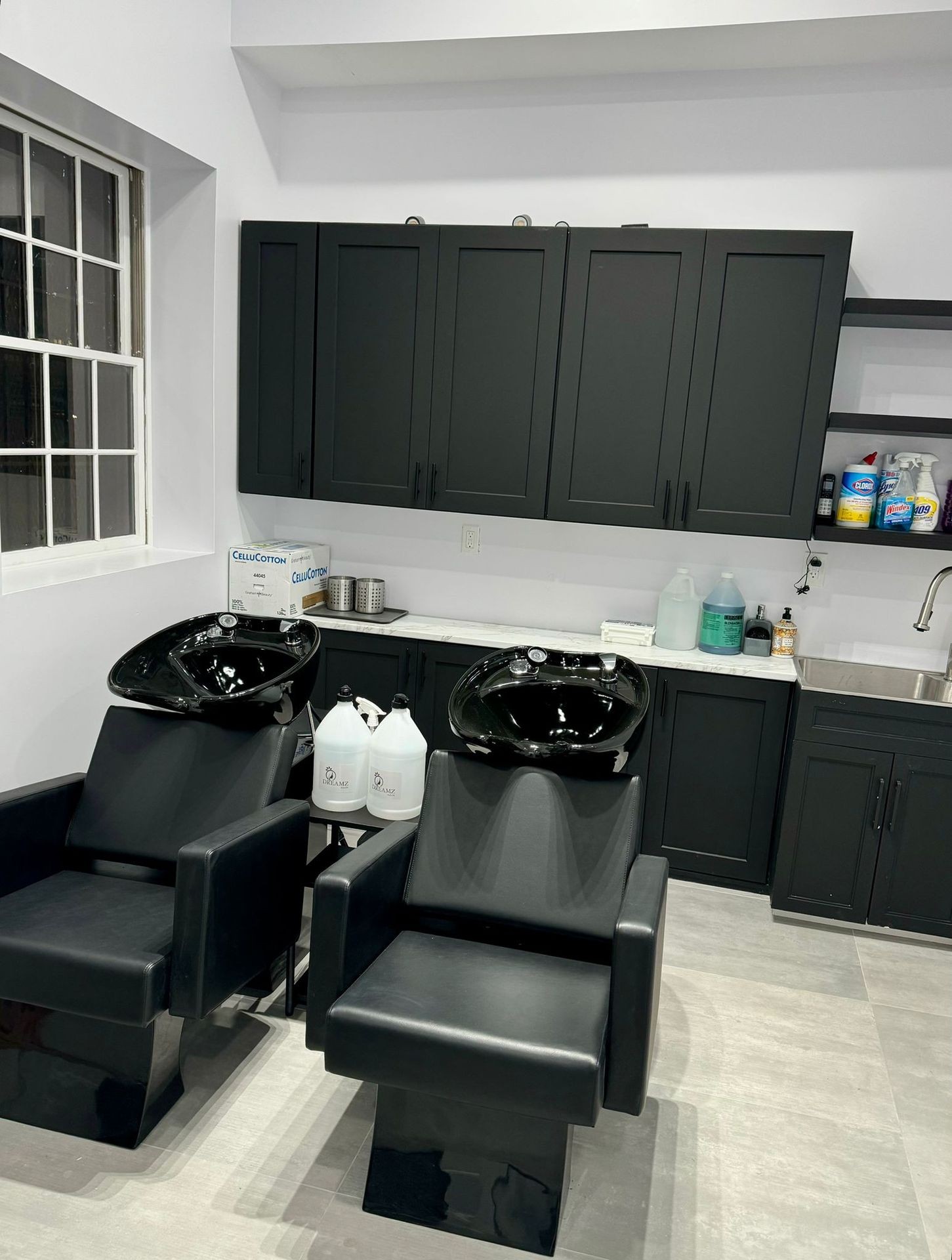 Hair salon wash station with black chairs and cabinets, bottles and cleaning supplies on countertops.